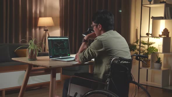 Asian Man Trader Sitting In A Wheelchair While Trading Cryptocurrency On Laptop Computer at Home