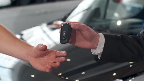 Young Man Giving Keys of Car To Buyer. Men Shaking Hands in Beautiful Car Dealership on Background