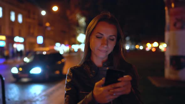 Attractive Woman Uses a Smartphone While Walking Through the Streets of the Evening City