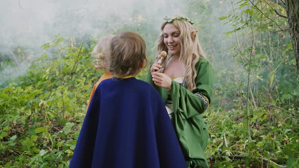 Fairy Elf Cosplay Young Woman Treats Little Babies Boys  with Candies in Green Forest