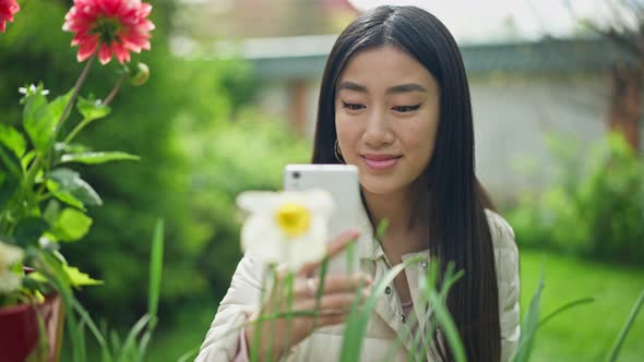 Proud Smiling Young Asian Florist Photographing Blooming Flowers on Smartphone in Garden