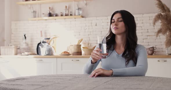 Young Thirsty Woman Holding Glass, Make Sip, Drinking Water, Sitting at Table in Home Kitchen