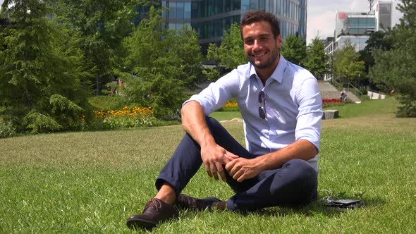 A Young Handsome Businessman Sits on Grass in a Park and Smiles at the Camera - Office Buildings