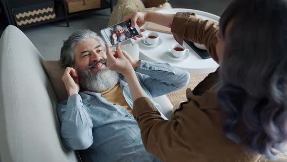 Joyful Mature Husband Posing for Smartphone Camera While Wife Taking Pictures Touching Screen