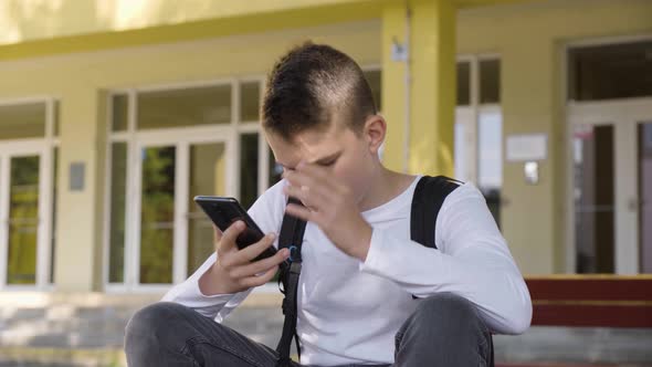 A Caucasian Teenage Boy Works on a Smartphone and Acts Frustrated As He Sits in Front of School