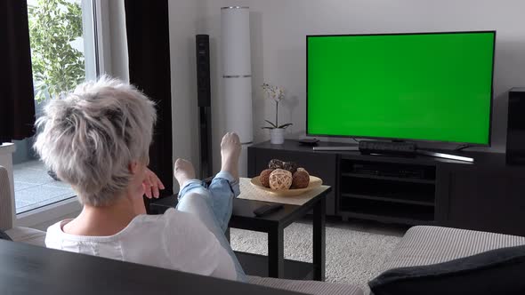 A Middle-aged Woman Sits on a Sofa in an Apartment Living Room and Watches TV with a Green Screen