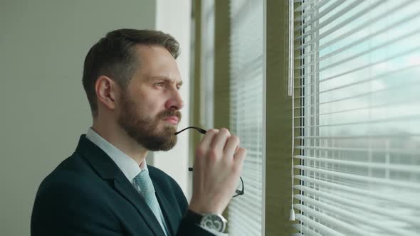 Thinking Process Focused Businessman in a Suit Stands at the Panoramic Window in the Office and