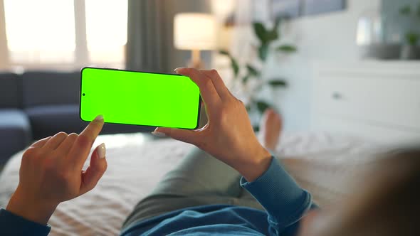 Woman at Home Lying on a Bed and Using Smartphone with Green Mockup Screen in Horizontal Mode