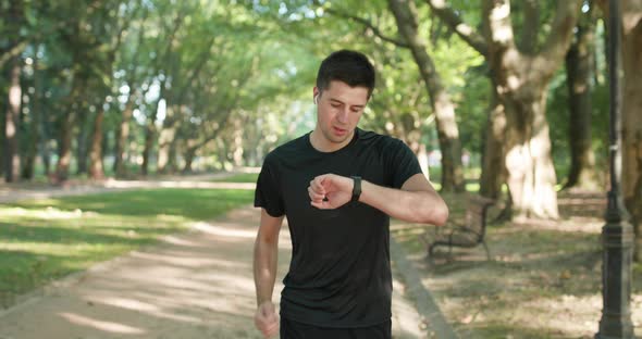 Sporty Man Running Along Empty Road in Park Checking Smartwatch Notification Online Using Earphones