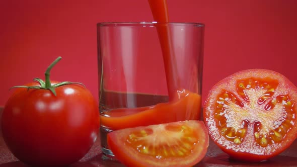 Closeup of Tomato Juice Poured in Glass Next to Tomato Halves on Red Background
