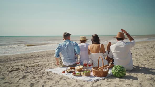 Slow Motion Rear View Of Four Happy Friends Sitting on Picnic Blanket Stands Up and Runs Along Sandy