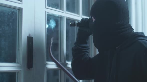 Close Up Of Thief Man Holding Flashlight And Crowbar Looking Inside The House While Standing Outside