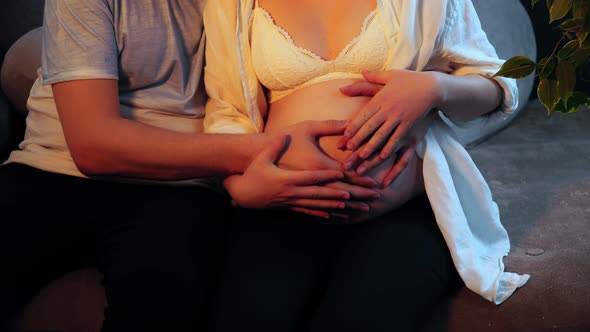 Pregnant Woman and a Man Touch Tenderly Belly and Hugging Sitting on a Sofa in Closeup