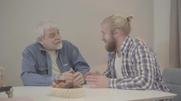 Adult Caucasian Son and Senior Father Laughing and Shaking Hands at Home. Young Man Telling Jokes