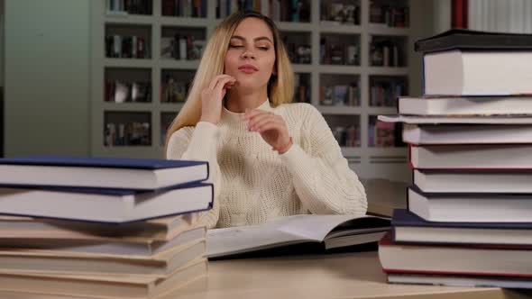 Tired Student Reads a Difficult Book in the Library Among a Large Pile of Books