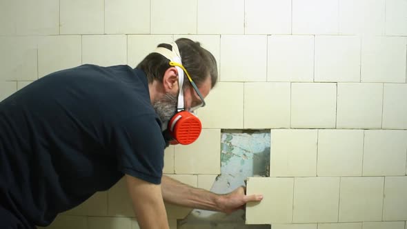 Bearded Worker in Respirator and Protective Glasses Dismantles By Hands an Old White Dirty Tile in a