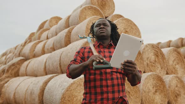 Young African Man, Farmer Holding Fork and Table in Front of the Hay Roll Stack. Smart Farming