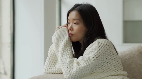 A Young Unhappy Asian Woman Sitting on the Couch with a Sad Face and Looking Out the Window