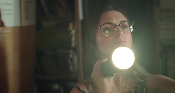 Serious Girl in Glasses with a Lantern Views the Library Shelves in the Darkness