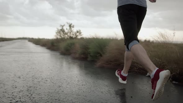 Legs of Man in Wet Sneakers Run Through Rain on Road Against Background of Cloudy Gray Sky and