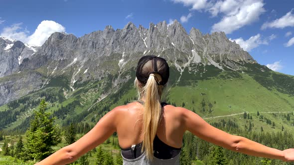 Pretty blond girl raising arms up in front of gigantic mountain landscape during summer - Rear close