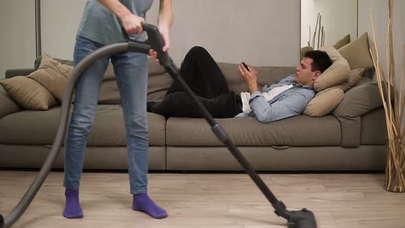 Unrecognizable Woman in Jeans Vacuuming Parquet Floor and Her Husband Plays on Smartphone on Couch