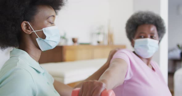 African american female physiotherapist wearing face mask helping senior female patient exercise