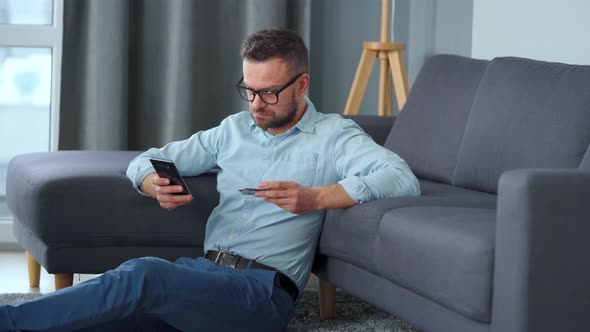 Bearded Man in Glasses Sits on the Floor Near the Sofa and Makes Payment Online with a Credit Card