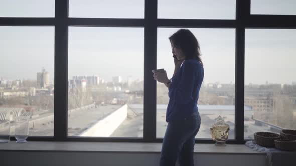 Side View of Brunette Caucasian Woman with Coffee Cup Standing in Front of Big Window Talking on the