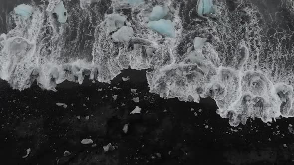 White water crashes over icebergs on black sand beach.