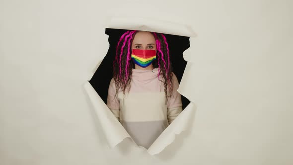 Young Woman in Protective Rainbow Mask Looking Out of Hole of White Background