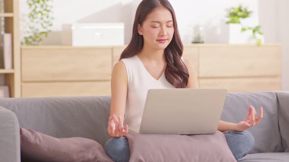 Clam of Asian young woman doing yoga lotus pose to meditation and relax on couch during work online