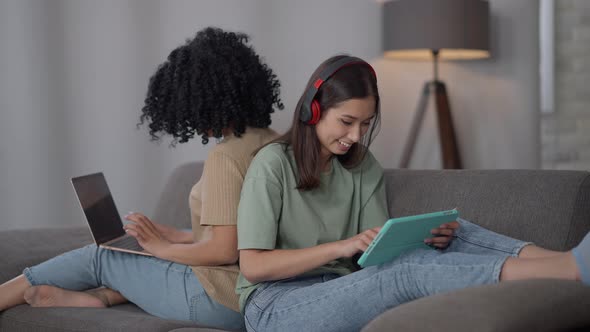 Joyful Slim Charming Asian Woman in Headphones Surfing Internet on Tablet As African American