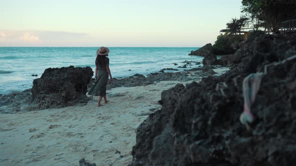 Woman is slowly walking on the beach near rocks on gently sea breeze, wearing a dress and sunhat. Sl