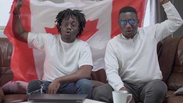 Portrait of Cheerful African American Men Shaking Canadian Flag Sitting Indoors on Couch Looking at