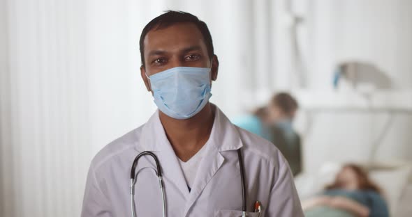 Mature Indian Doctor Wearing Face Mask Standing in Hospital Ward Talking at Camera