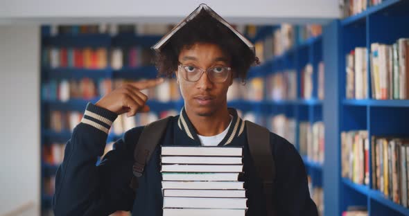 Young African Student Looking in Camera with Book on Head in Library of University