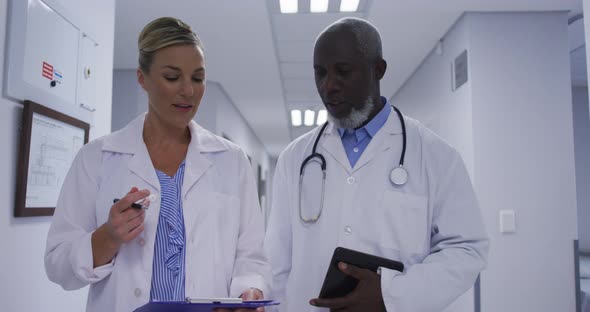 Diverse couple of male and female doctors walking hospital corridor and looking at tablet