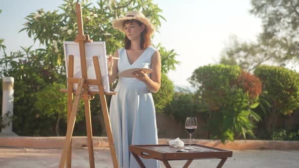 Portrait of Concentrated Positive Caucasian Woman in Dress and Straw Hat Painting in Slow Motion