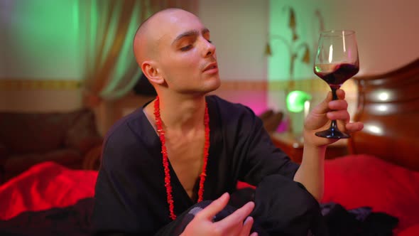 Confident Male Queer Shaking Red Wine in Glass Looking at Camera Sitting in Bedroom with Red Light