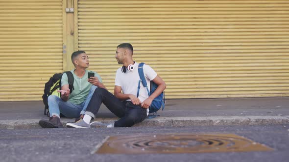 Two happy mixed race male friends sitting, using smartphone in the street