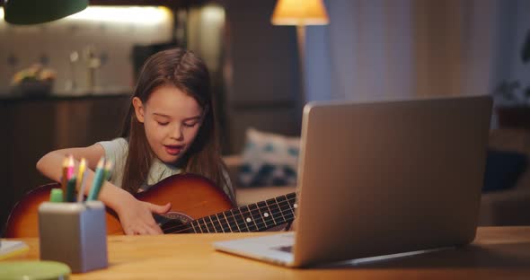 Happy Little Child Learning To Play Guitar While Watching Lessons at Laptop and Smiling at Home