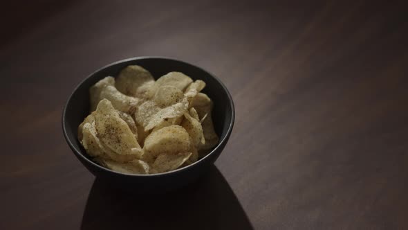 Slow Motion Handheld Shot of Organic Potato Chips with Black Pepper in Black Bowl on Walnut Table