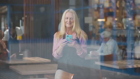 View Through Window of Smiling Aged Waitress in Apron Using Smartphone Standing in Cafe