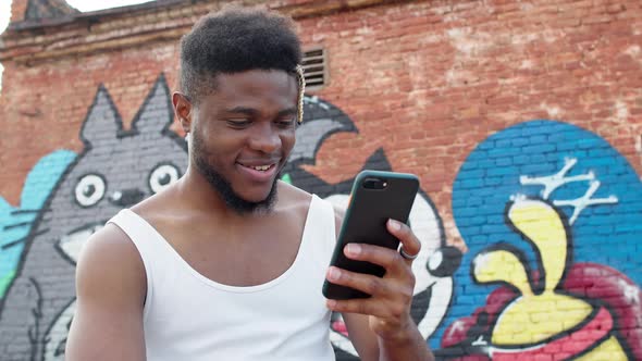 Young Black Guy With Mobile Phone In His Hands. He On Graffiti Wall Background.