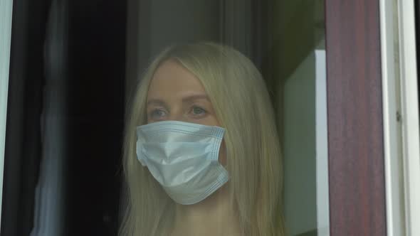 A young woman in a medical protective mask looks out the window