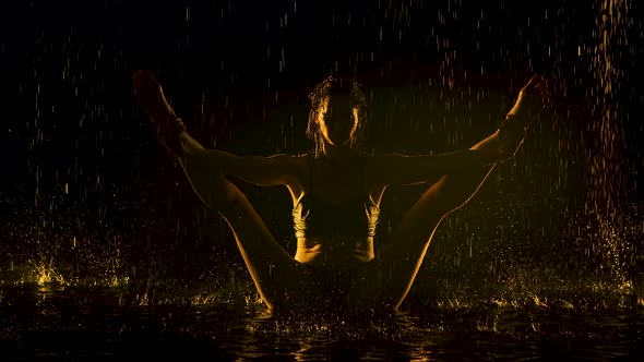 A Young Woman Balance with Legs Wide Apart in a Dark Studio Under the Rain. A Slim Body Silhouetted