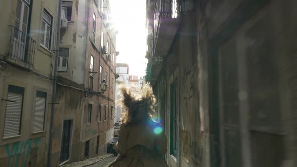A Young Stylish Woman Runs Down a Narrow European Street with Her Hair Flowing in Front of the Sun