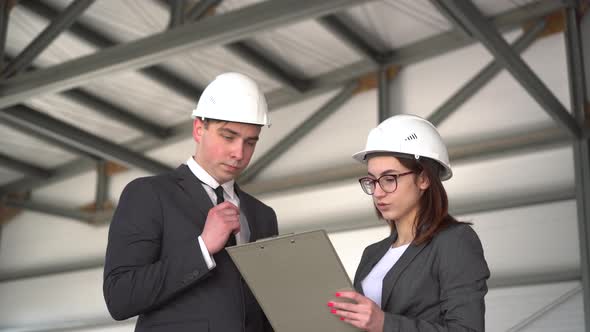 Young Man and Woman in Helmets with Documents at a Construction Site. Chiefs in Suits Discussing the