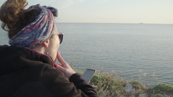 A woman smoking cigarette and playing with phone on the hill with a sea view.